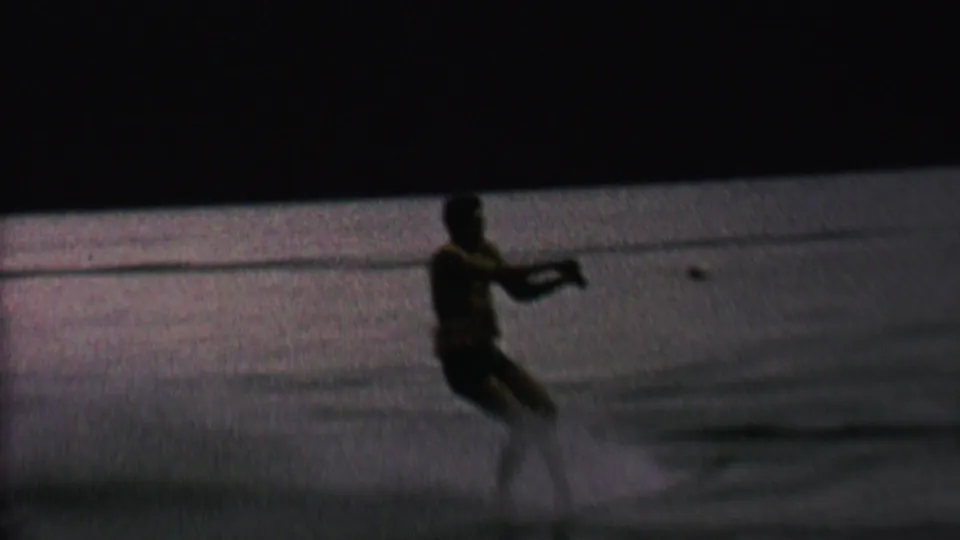A male water skier in a dark swimsuit holds the tow rope while gliding across the lake in grainy 1962 home-movie footage.