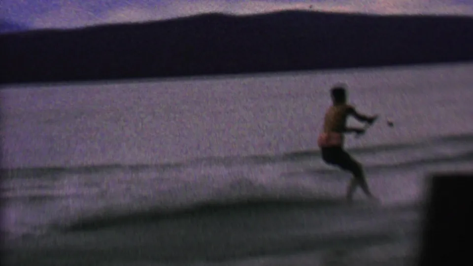 The water skier leans back with knees bent as the tow line pulls him across the lake, with heavy grain and blur from the 8mm home-movie scan.