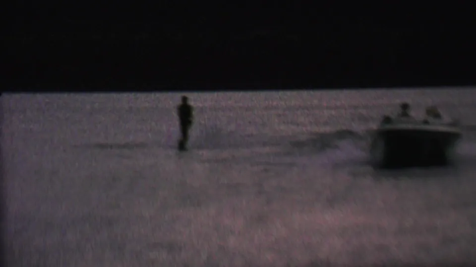 A small motorboat with passengers pulls a distant water skier across the lake, with the boat wake visible in the low-light 1962 home-movie frame.