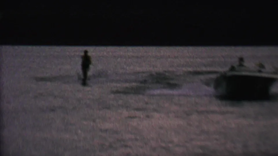 A distant man water skis on a lake near a small boat in supplied 1962 St. Paul, Minnesota home-movie footage, seen in grainy low evening light.