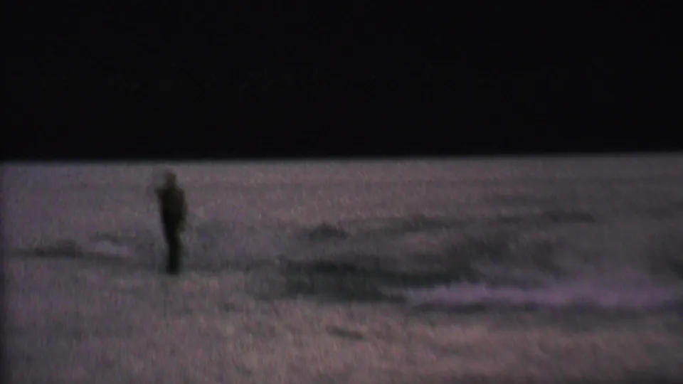 A silhouetted skier moves across the lake surface in low-light 8mm footage, with the shoreline reduced to a dark band in the background.