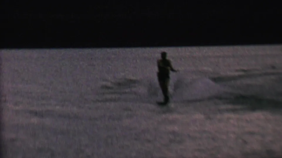 The skier remains upright on the water, photographed as a small dark figure against the textured lake surface in vintage 8mm home-movie footage.