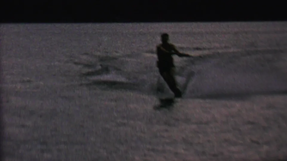 A man on water skis leans with arms extended while moving across the lake, appearing as a dark silhouette in the supplied 1962 home-movie scene.