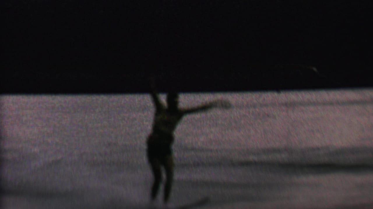 The water skier, seen in low light on the lake, appears to lose balance while one arm lifts and the tow rope angles across the frame.