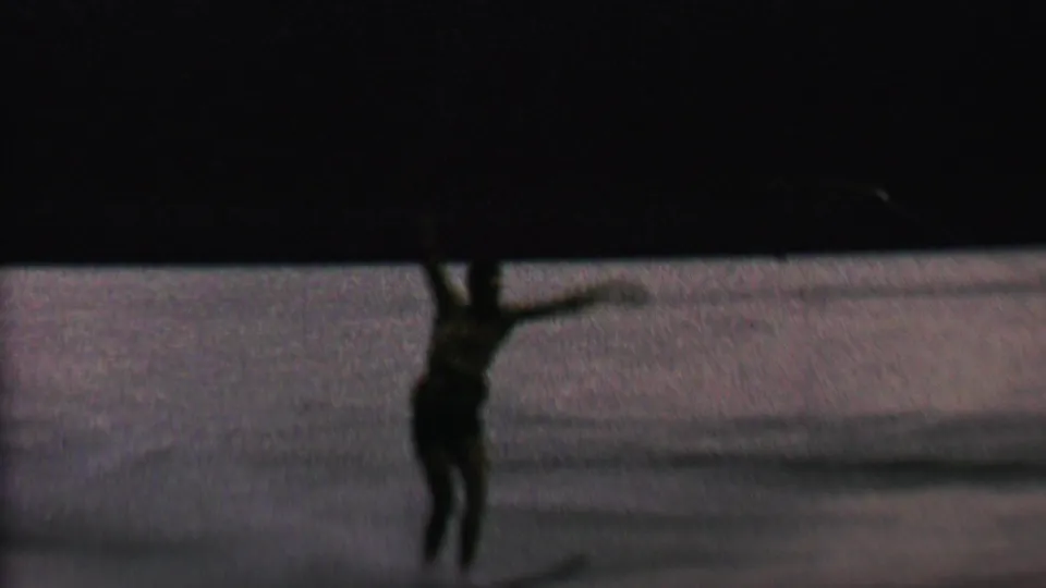 The water skier, seen in low light on the lake, appears to lose balance while one arm lifts and the tow rope angles across the frame.