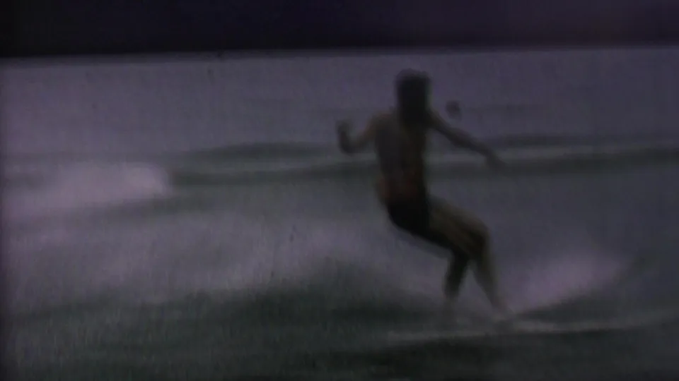 The skier leans backward near the water surface, with the rope and skis still visible as the fall begins in the grainy 8mm frame.