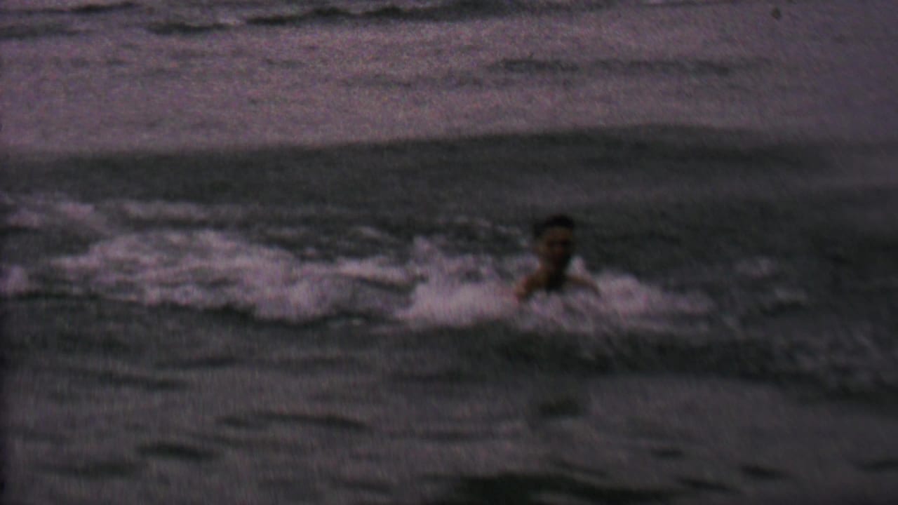 A man appears in choppy lake water in 1962 home-movie footage, with a small board or floating object nearby and no clear shoreline detail visible.