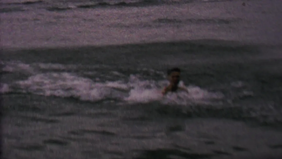 A man appears in choppy lake water in 1962 home-movie footage, with a small board or floating object nearby and no clear shoreline detail visible.
