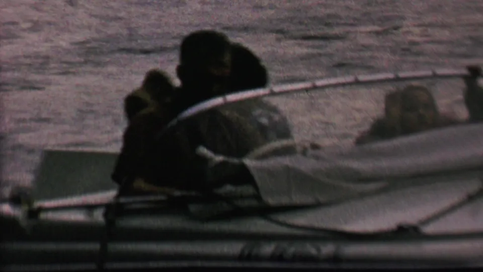 Several people sit close together in a small motorboat on a lake, including one person in a dark hat, in grainy 1962 home-movie footage.
