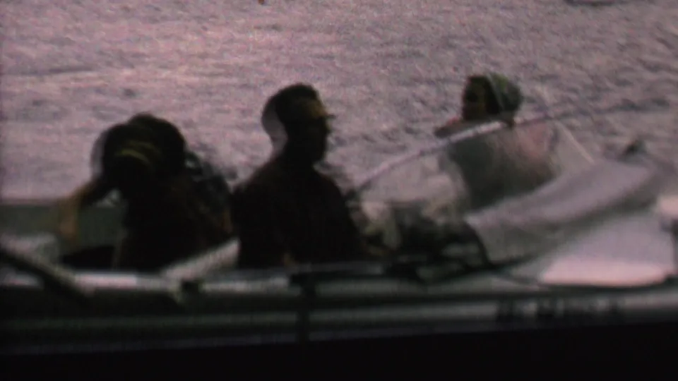 Three people are visible aboard a small motorboat on dark lake water, with one figure leaning forward and another seated near the stern in the 1962 home-movie scene.
