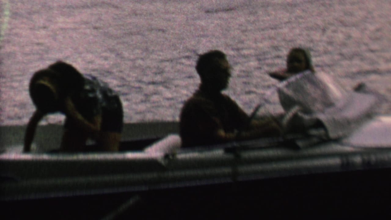 1962 home-movie frame from metadata showing a small family motorboat on water, with seated passengers partly silhouetted by backlight and soft 8mm focus.