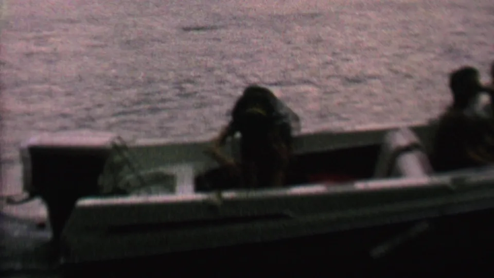 Backlit 8mm frame of passengers in small open boats on a lake, with one person leaning forward and another seated near the stern area.