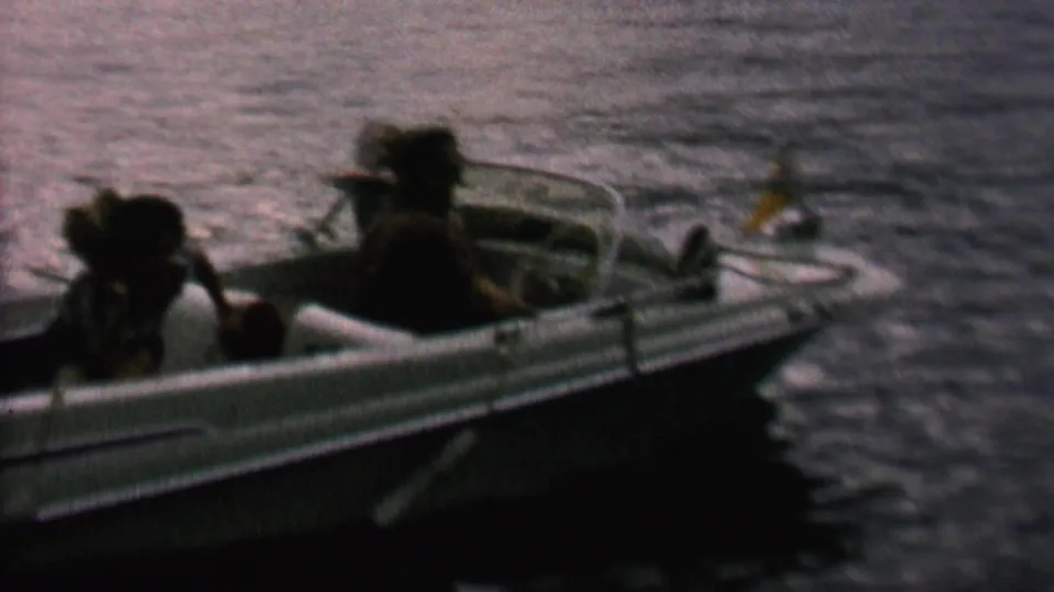 Home-movie frame showing a small motorboat moving across water, with several passengers seated low and the boat edge cutting through the lower frame.