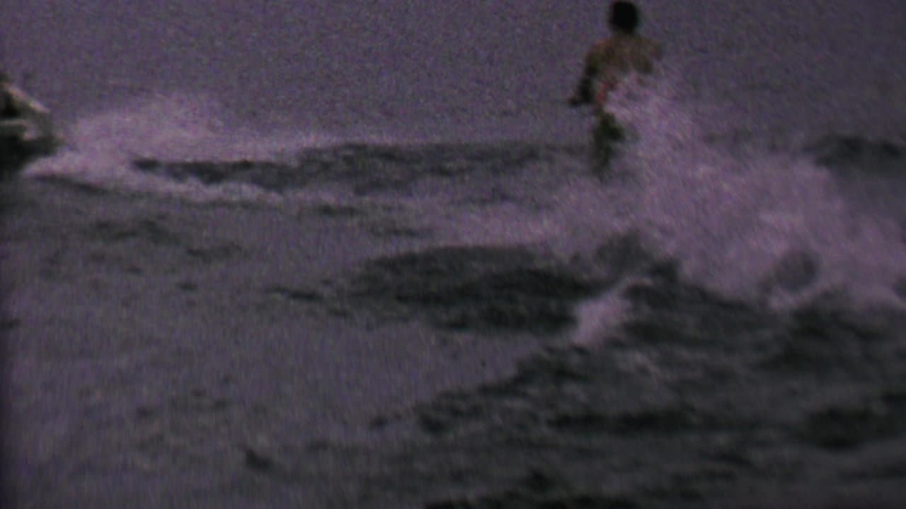 A distant rope-towed waterskier rides over choppy water in the supplied 1962 St. Paul, Minnesota home-movie clip, with heavy spray and low-light grain.