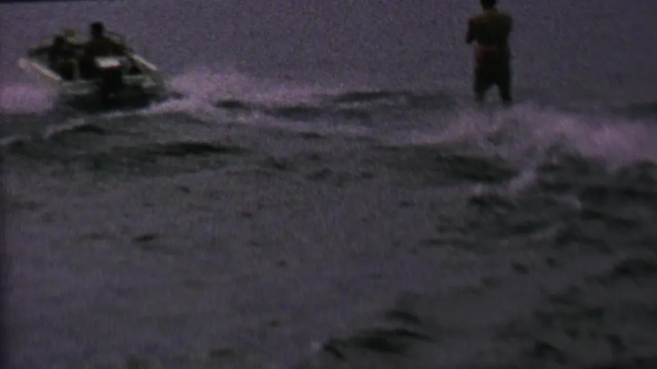 A small motorboat and a standing waterskier are visible on a dark lake in this archival 8mm frame, consistent with the supplied 1962 St. Paul waterskiing metadata.
