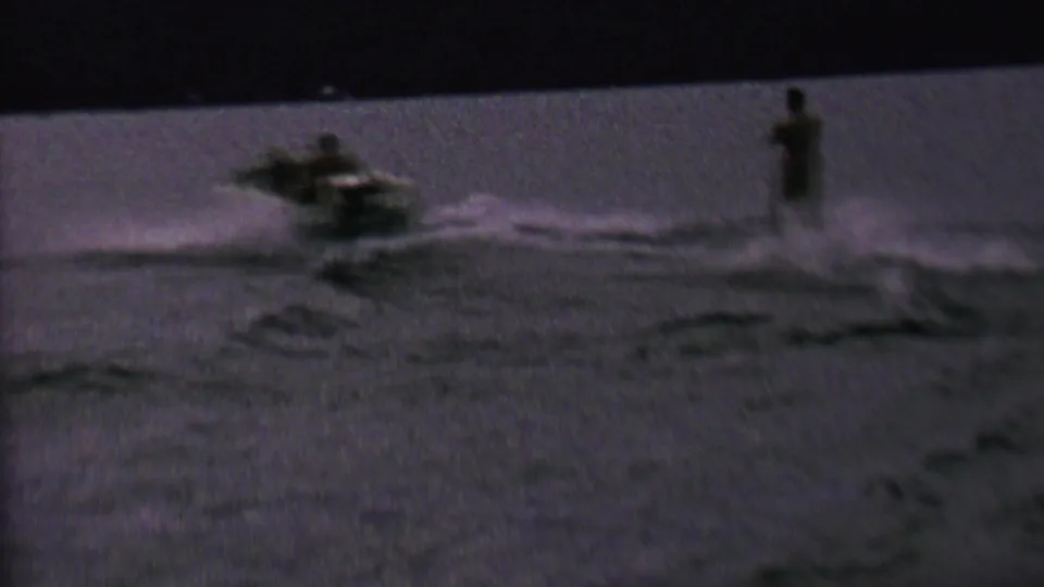 In 1962 home-movie footage from the St. Paul, Minnesota area, a small motorboat pulls a distant water skier across a dark, grainy lake surface.
