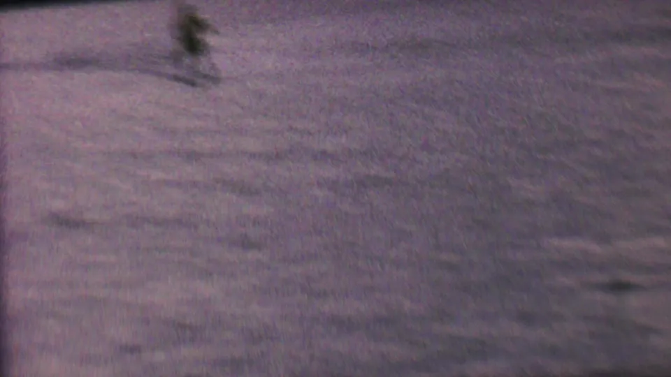 The skier appears as a small blurred figure on the lake, with the tow action moving farther from the camera in purple-tinted 8mm footage.