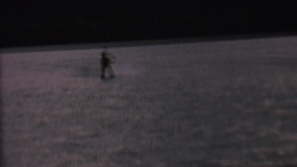 A small, distant skier remains visible against the dark lake surface as the home-movie shot softens and the action recedes.
