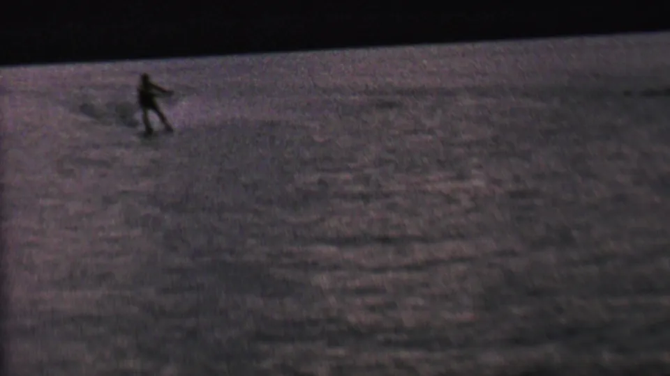 A small silhouetted water skier glides over rippled lake water in soft, purple-tinted 8mm home-movie footage.