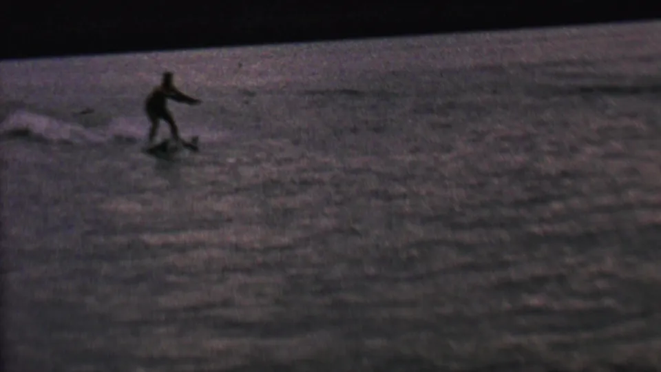 A water skier appears near the left side of the frame, riding over dark lake water in grainy 8mm home-movie footage.