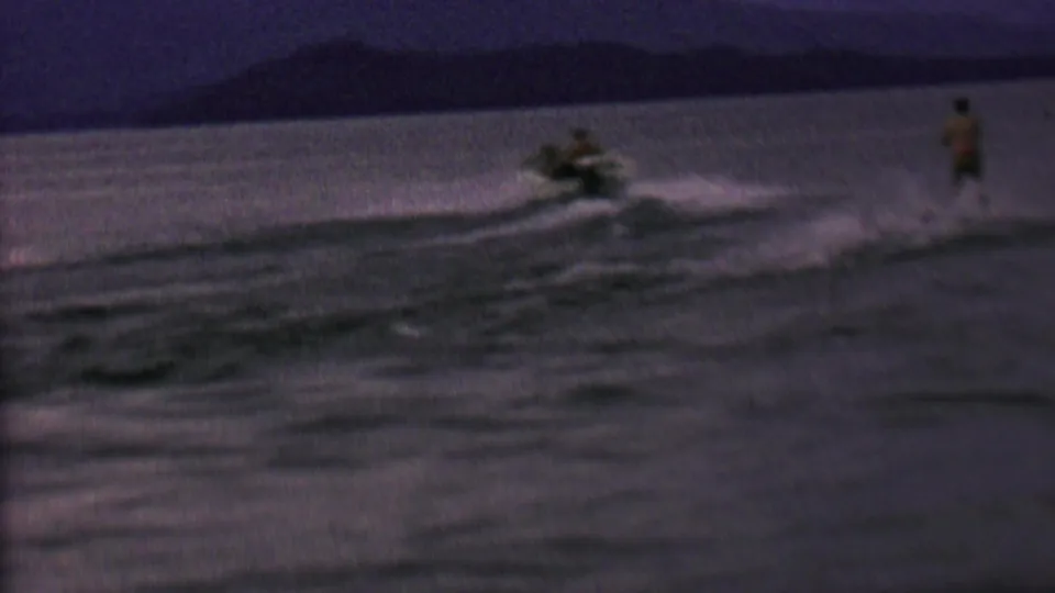 The motorboat and water skier are seen farther apart across the lake, with dark open water and a distant shoreline in this short 1962 8mm home-movie view.