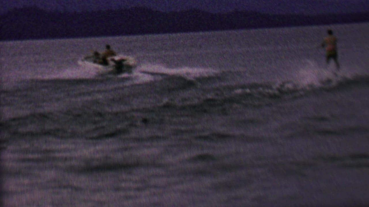 In this 1962 United States home-movie frame, a small motorboat crosses dark lake water while a distant person appears to water ski behind it.