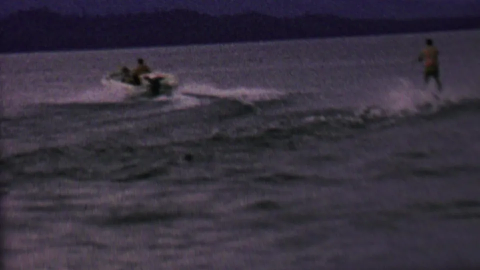 In this 1962 United States home-movie frame, a small motorboat crosses dark lake water while a distant person appears to water ski behind it.