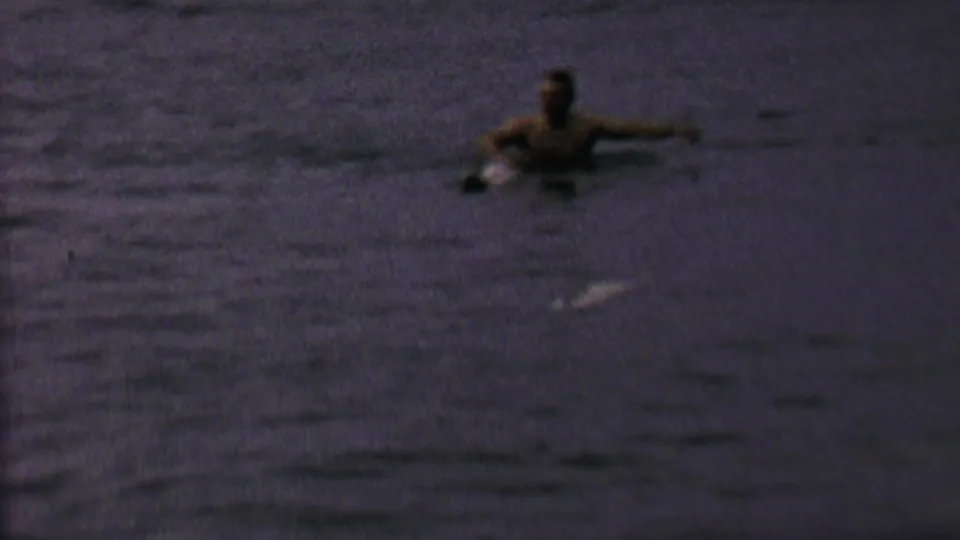 A person appears to float or swim in dark lake water in the 1962 home-movie footage, with the shore faintly visible in the background.