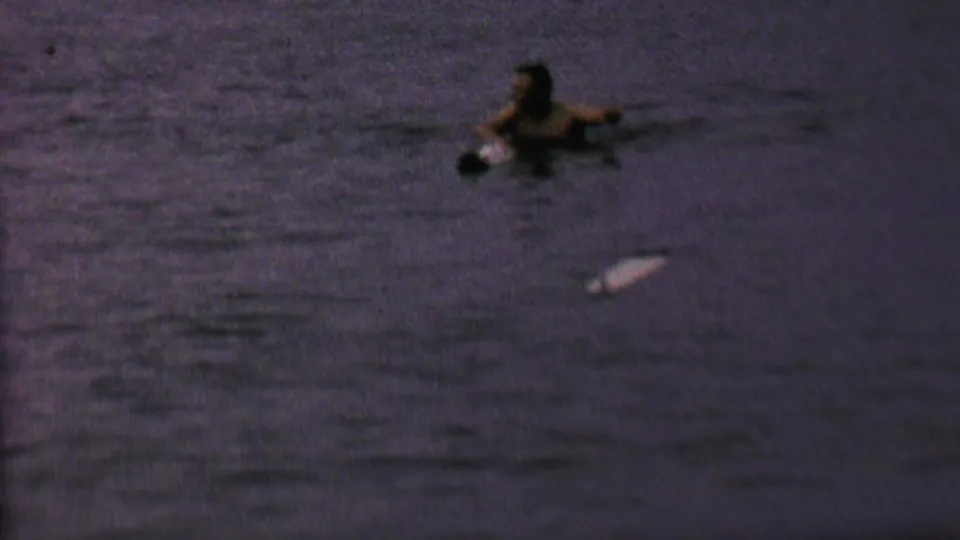 A swimmer is seen in dark lake water near a small light-colored object, with the frame retaining the grain and purple cast of 1962 8mm footage.