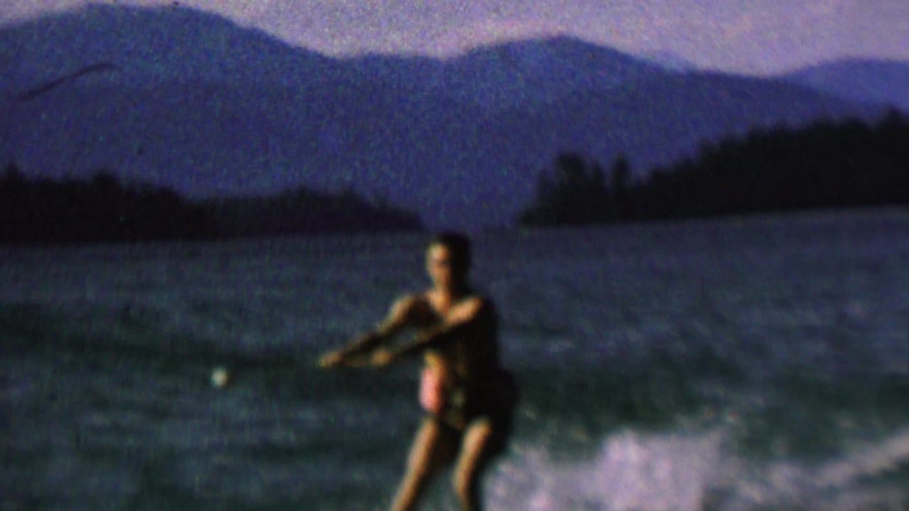 A male water-skier appears on a lake in archival 1962 United States home-movie footage, holding a tow rope as spray rises behind him.