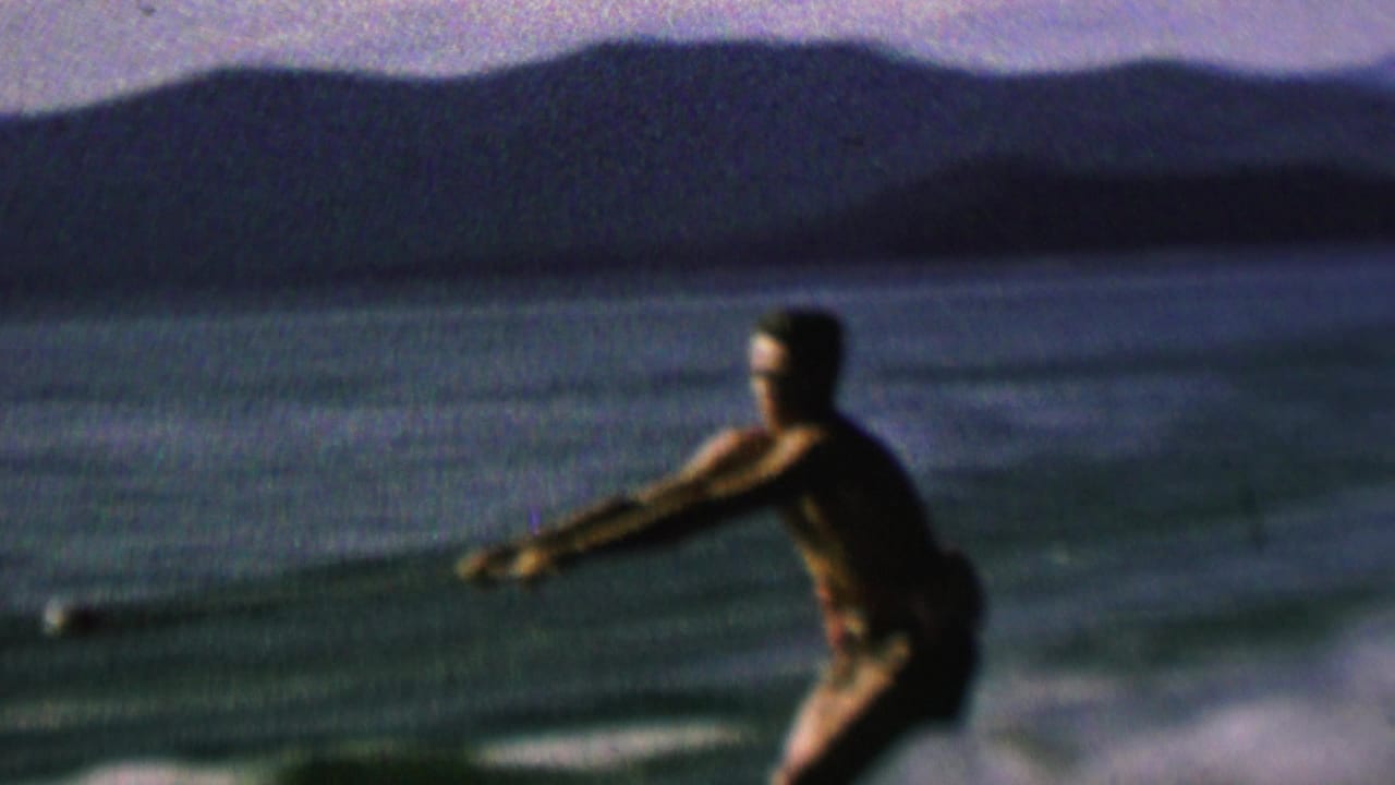 A man water-skis across open water, leaning into the pull of the rope with a shadowed shoreline and distant hills visible beyond the lake.