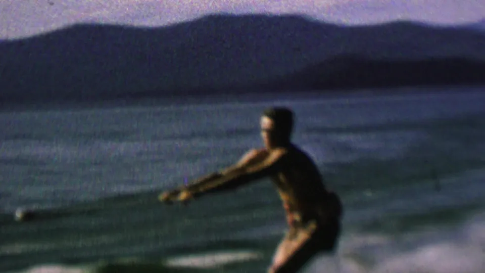 A man water-skis across open water, leaning into the pull of the rope with a shadowed shoreline and distant hills visible beyond the lake.