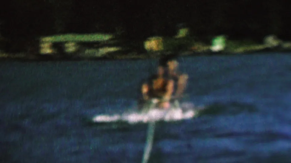 A male water-skier moves through a bright splash of wake on the lake, with the rope extending forward and the distant shoreline behind him.
