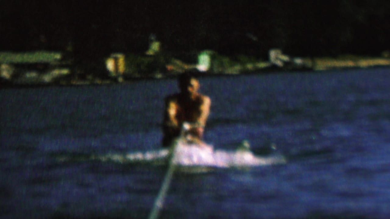 In supplied 1962 St. Paul home-movie footage, a male water skier rides low on dark lake water while holding a tow rope, with spray behind him and a dark shoreline in the background.