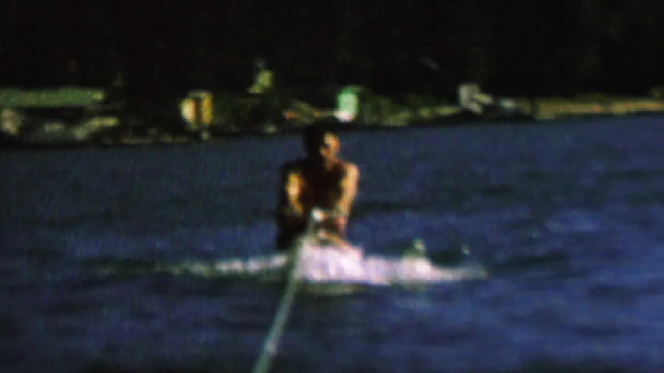 In supplied 1962 St. Paul home-movie footage, a male water skier rides low on dark lake water while holding a tow rope, with spray behind him and a dark shoreline in the background.