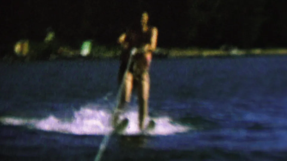 A male water skier stands upright behind the tow rope on a dark blue lake, with white wake under the skis and a mostly black tree line beyond the water.