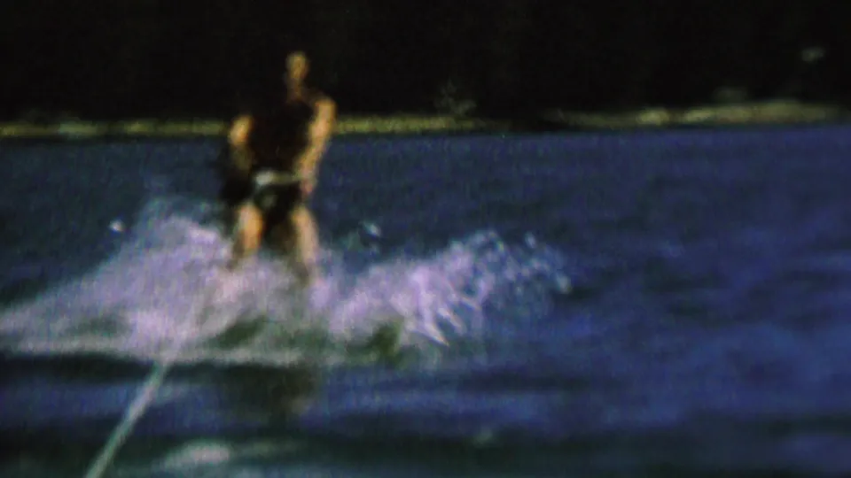 The skier moves closer in the frame, holding the tow rope as white spray spreads across the dark lake surface under low artificial light.