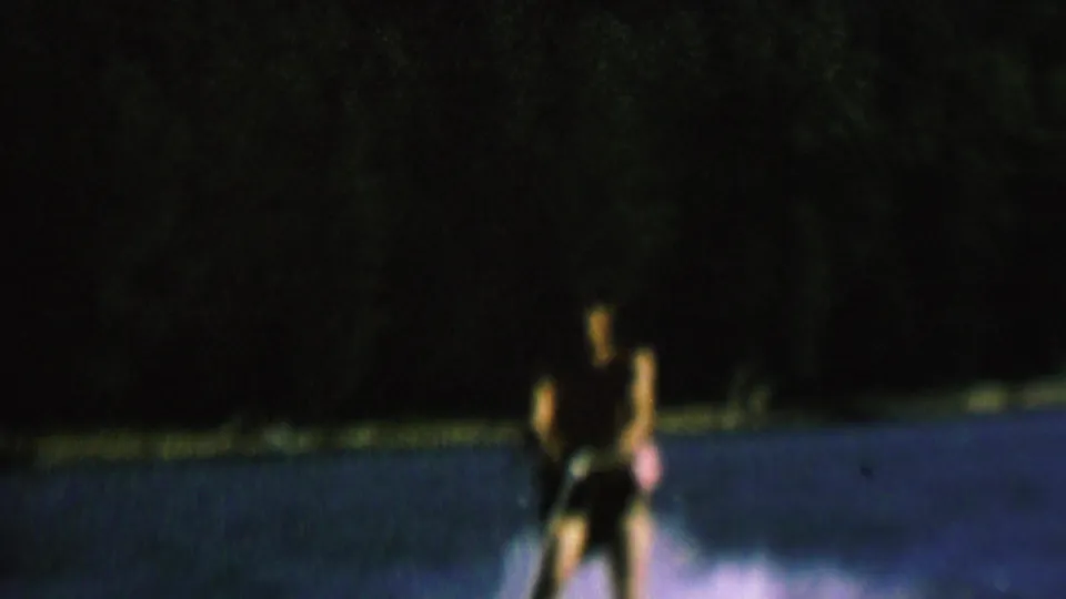 The water skier appears farther away on the lake, still holding the tow rope, with a small patch of wake and a dark wooded shoreline filling much of the night frame.