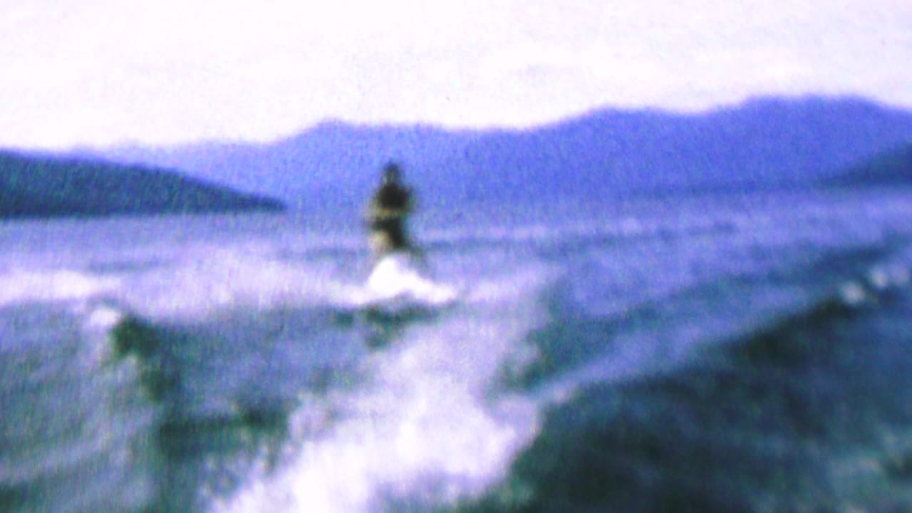 A water skier appears on a lake in grainy 1960s 8mm footage, with white wake spray behind them and a hazy mountain or ridge line across the background.