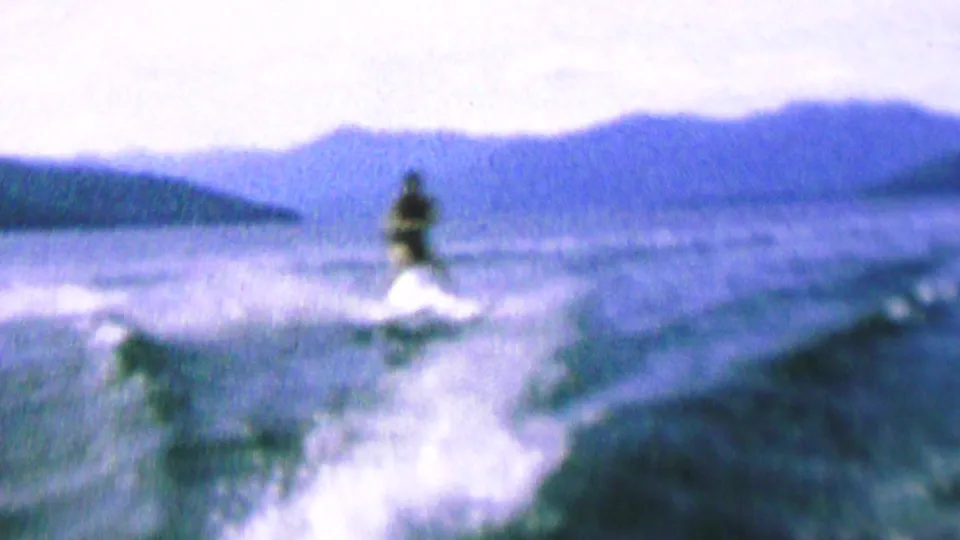 A water skier appears on a lake in grainy 1960s 8mm footage, with white wake spray behind them and a hazy mountain or ridge line across the background.