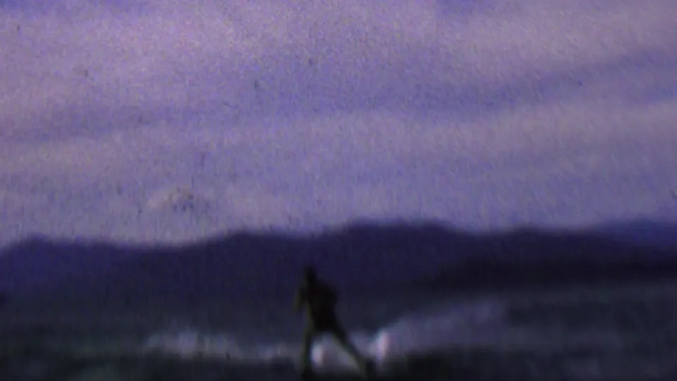 The skier is seen as a dark silhouette against low evening light, crossing the lake with blurred mountains or ridges and wake spray behind them.