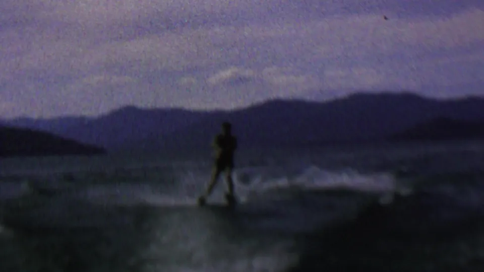 A dark figure on water skis moves across the foreground of the lake, with wake spray, a shadowed shoreline, and layered ridge shapes in the background.