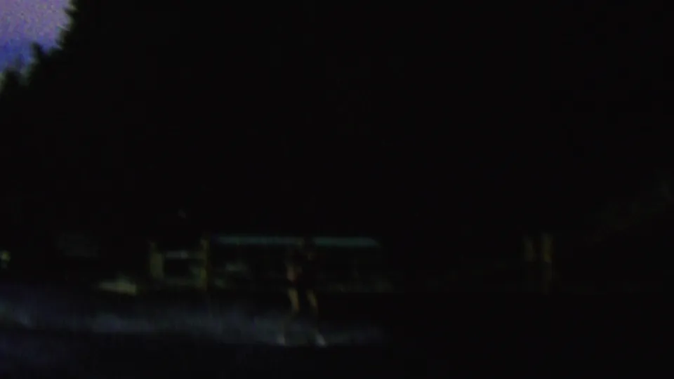 The final frame becomes very underexposed, showing a dark shoreline or hillside with faint horizontal dock-like shapes near the water and dim ridge silhouettes beyond.