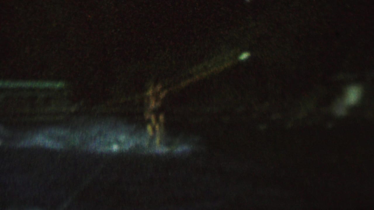 In dark 1962 home-movie footage, a distant female water skier appears on a lake surface with a pale tow rope and faint spray visible in the spotlight.
