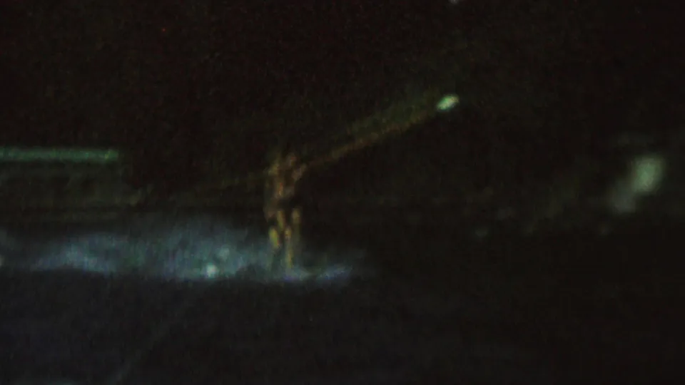In dark 1962 home-movie footage, a distant female water skier appears on a lake surface with a pale tow rope and faint spray visible in the spotlight.