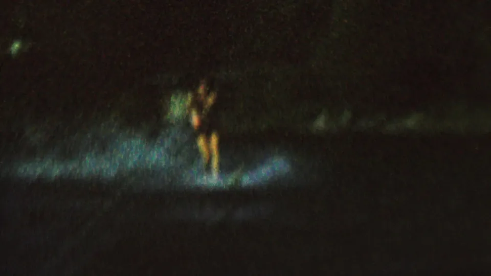 A female water skier glides through a blue-white spotlight on the dark lake, with motion blur and heavy 8mm grain obscuring fine detail.