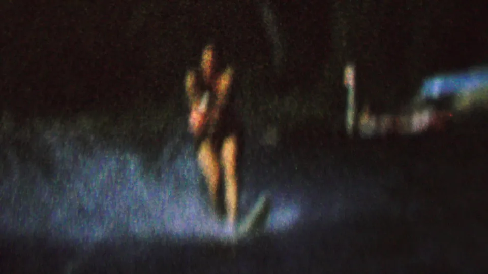 A woman on water skis moves across a night lake, with blue-white light catching the spray and trees barely visible behind her.