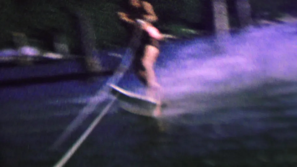 The skier is seen closer to the camera, leaning while holding the tow rope as the ski cuts through dark water and throws a blue-toned spray.