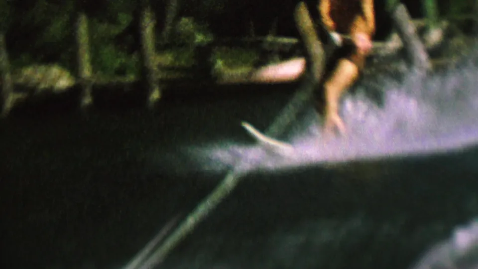 A woman water skier passes near the filming boat, holding the tow rope as a large spray arcs from the ski against the dark lakeshore.