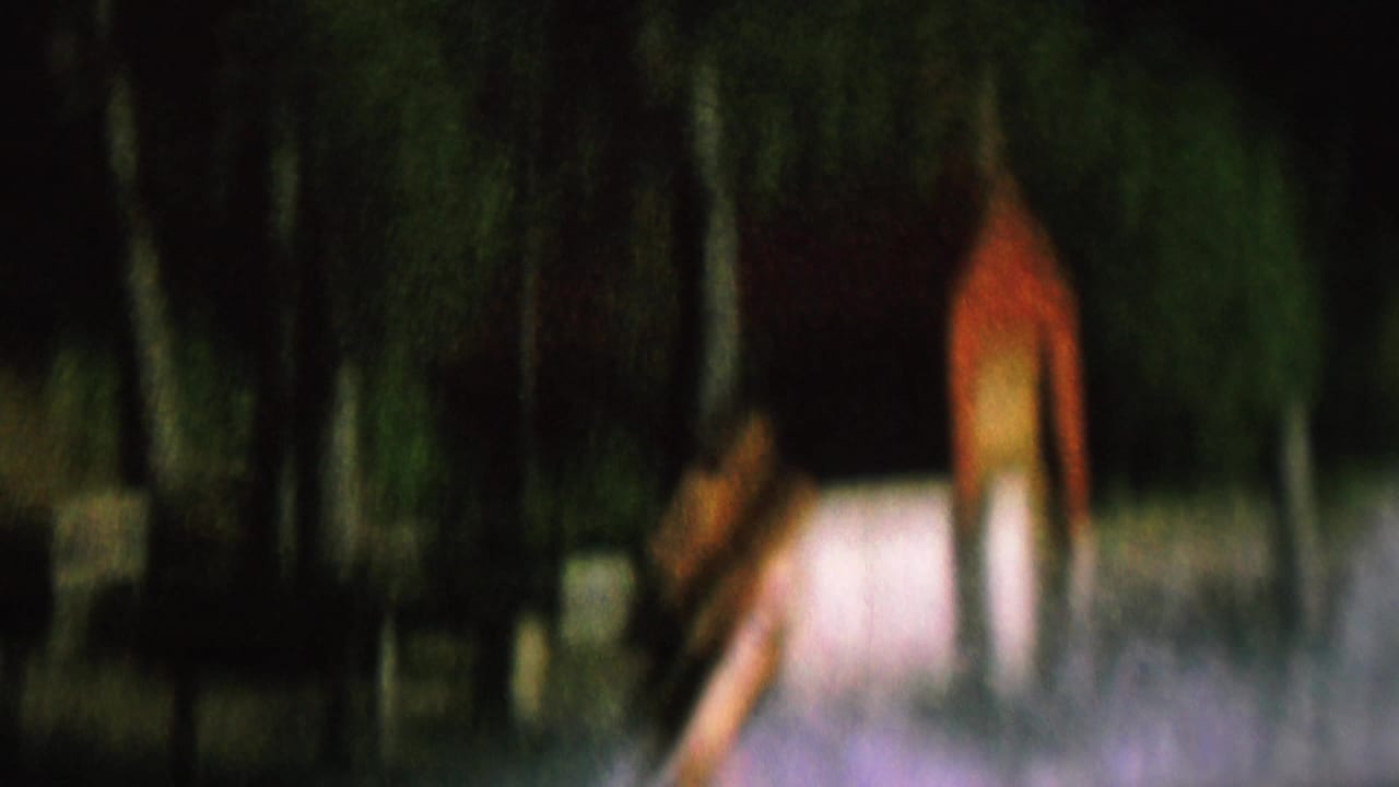 1962 home-movie frame from the United States showing a distant water skier on a lake near trees and indistinct shoreline structures.